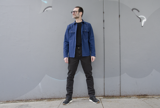 matt standing in front of a grey wall with overdye black skinny jeans and blue wander overshirt